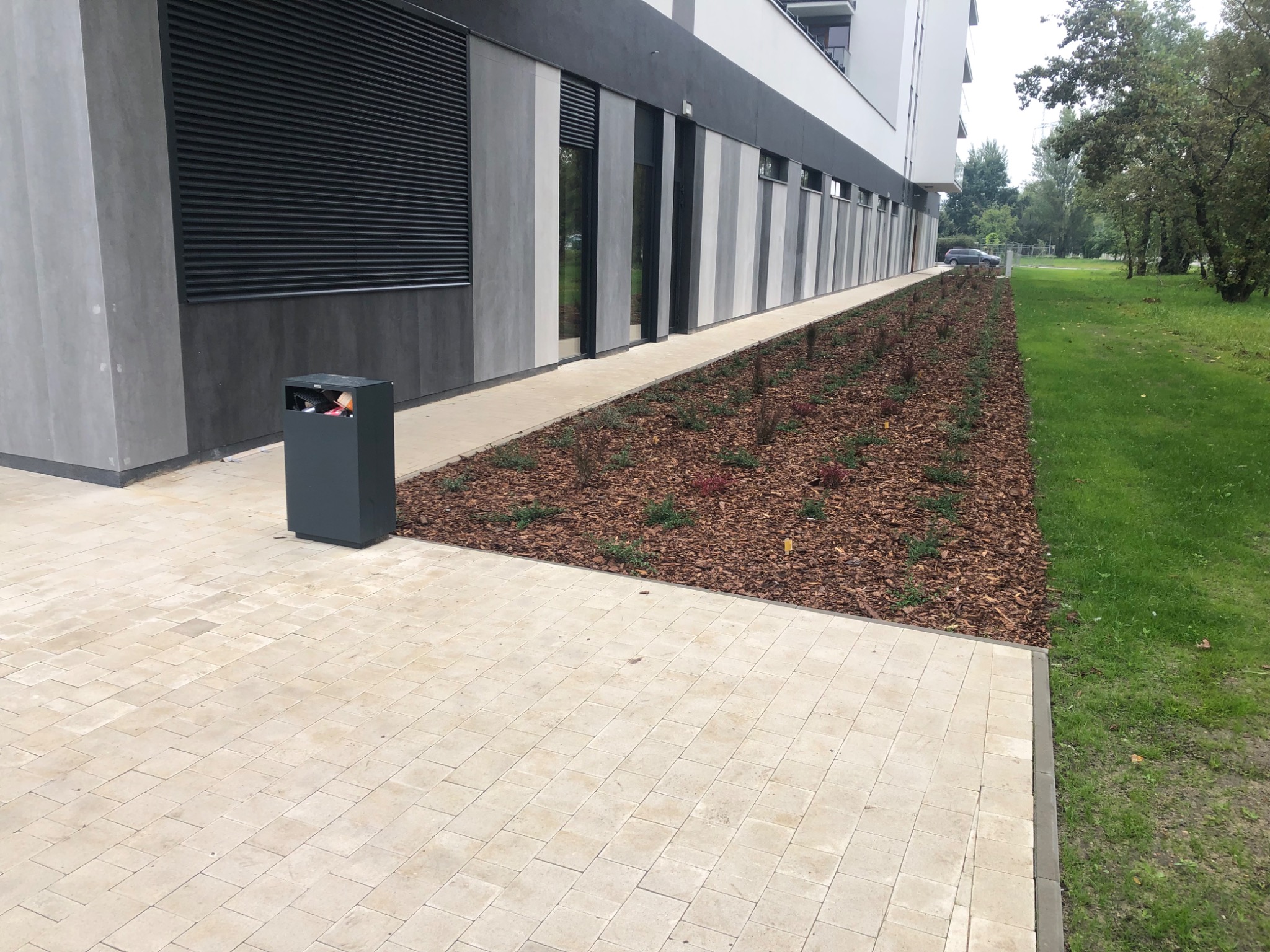 Beżowa kostka brukowa ułożona przed nowoczesnym budynkiem z elementami szarości, obok rabata z korą i młodymi roślinami, widoczny kosz na śmieci.
