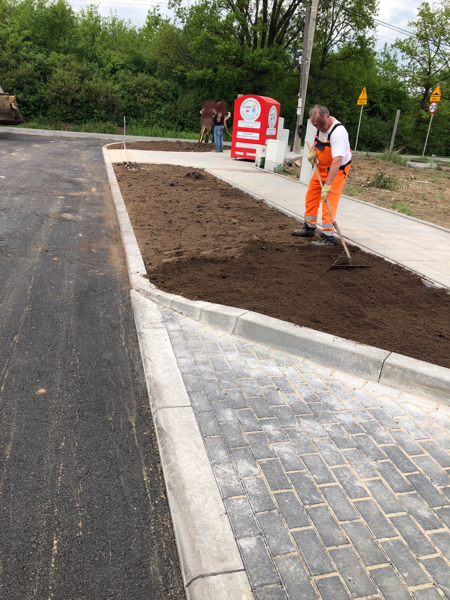 Pracownik w pomarańczowym kombinezonie wyrównuje ziemię grabiami na przygotowanym terenie obok nowej drogi asfaltowej i chodnika z kostki brukowej.
