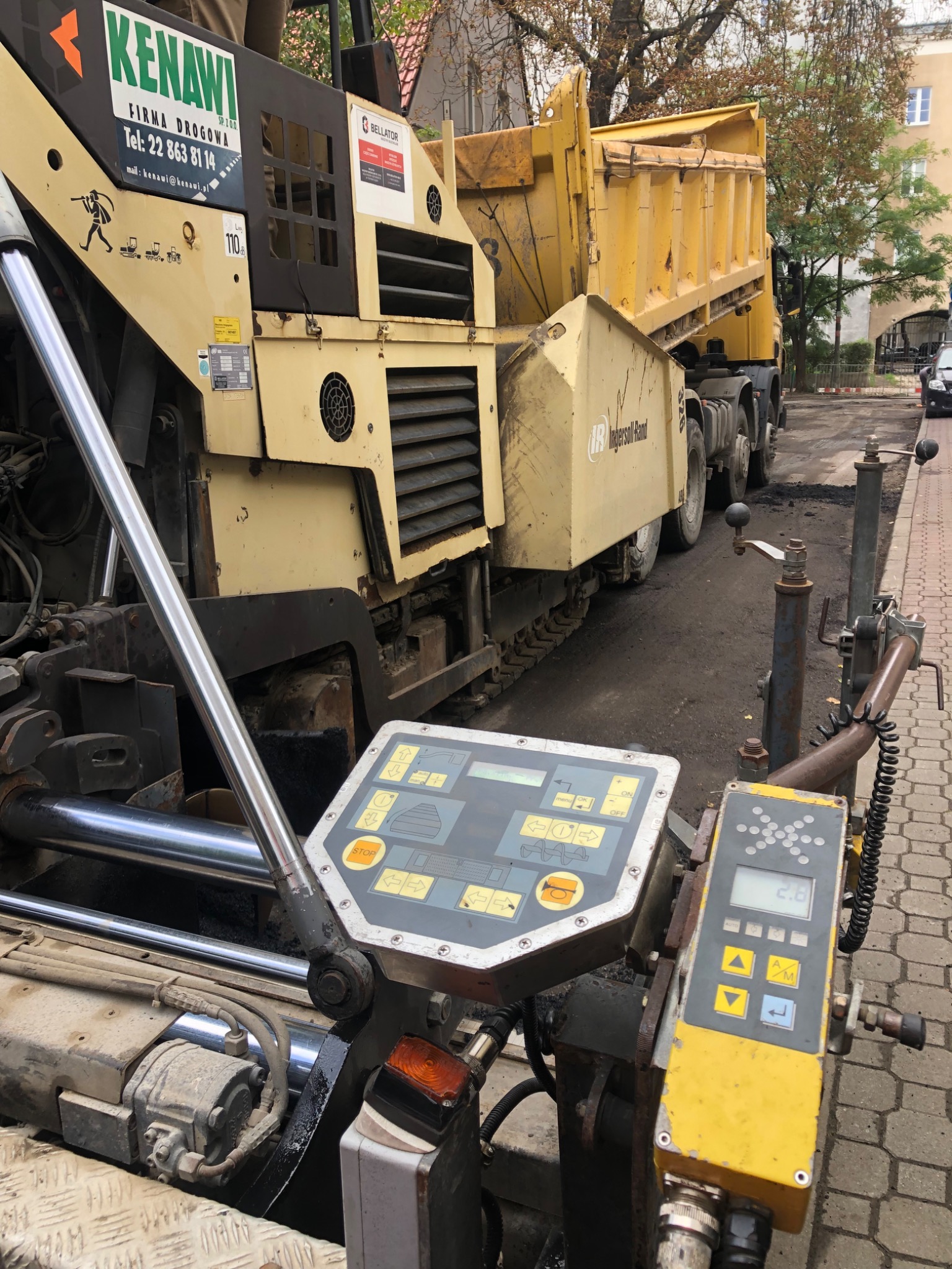 Rozściełacz asfaltu marki Kenawi z podłączoną wywrotką Ingersoll Rand na tle miejskiej ulicy w trakcie robót drogowych.