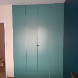 Czterodrzwiowa szafa w kolorze morskim, wkomponowana w narożnik pokoju z dwiema różnymi kolorami ścian, widoczna z perspektywy niskiego kąta.