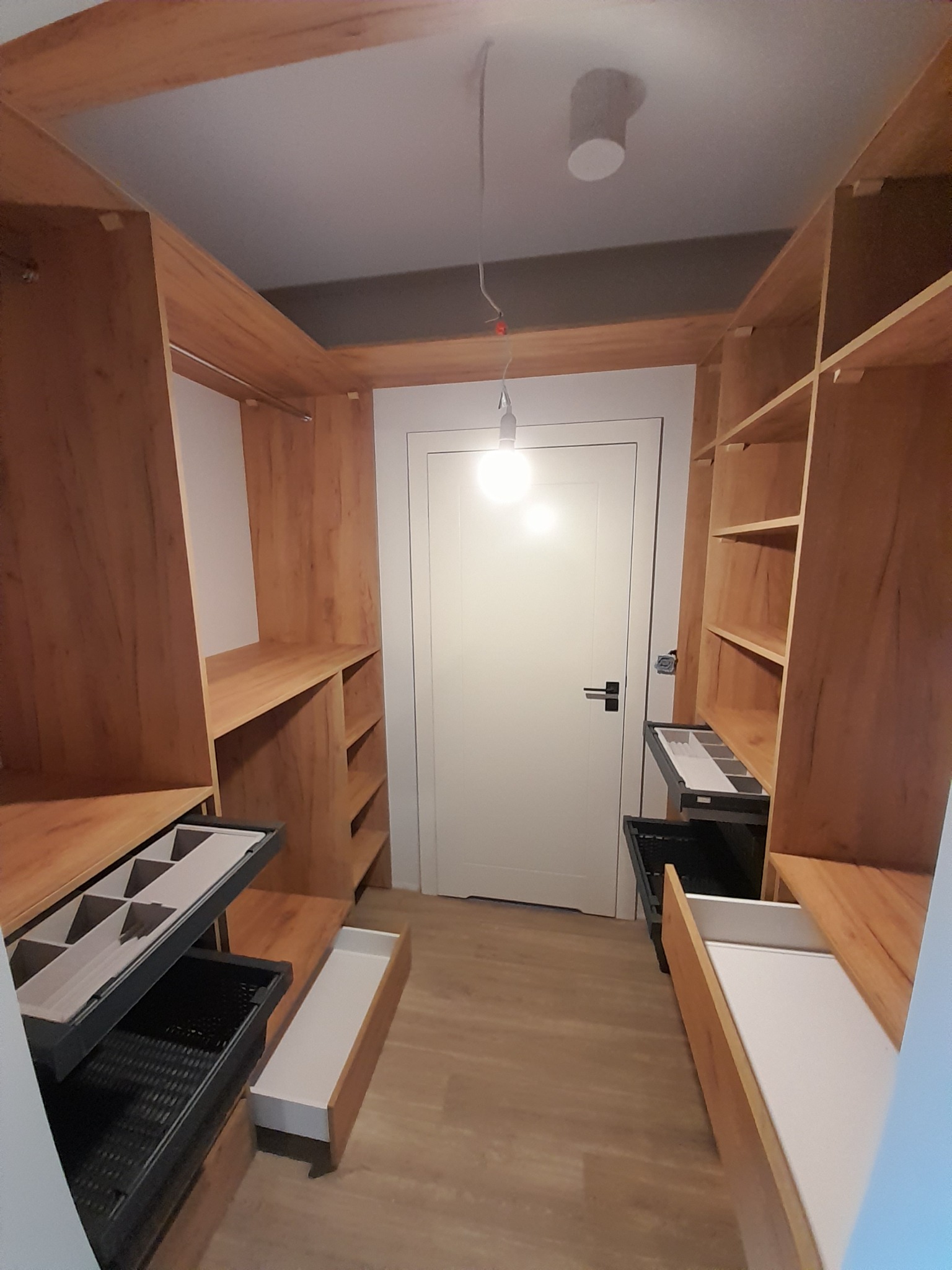 Wnętrze szafy wnękowej z drewnianymi półkami i wysuwanymi szufladami, widok z perspektywy wejścia, oświetlenie sufitowe.