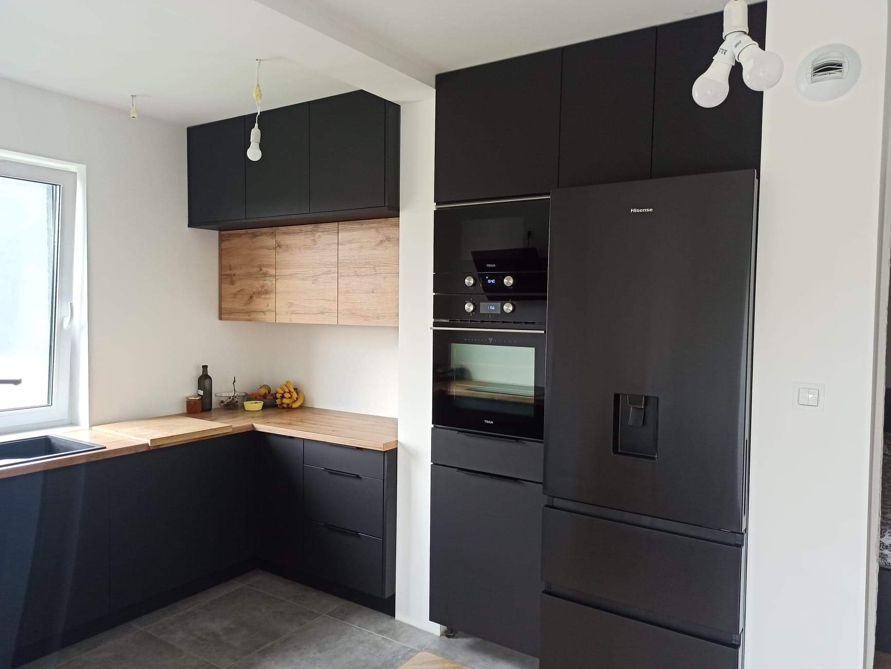 Nowoczesna kuchnia z czarnymi szafkami, drewnianymi blatami i sprzętem AGD w zabudowie, oświetlona wiszącymi żarówkami.