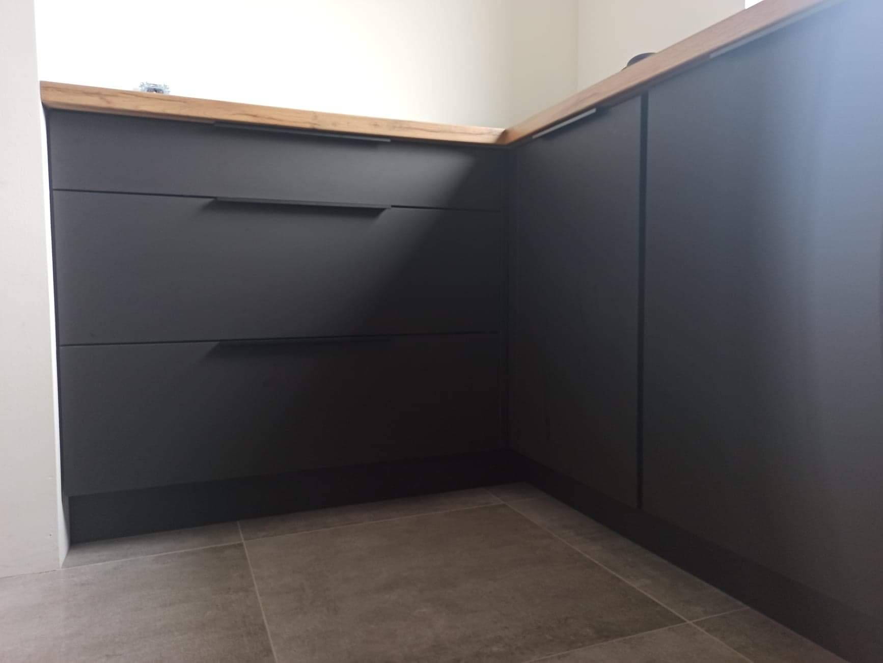 Fragment zabudowy kuchennej z ciemnymi frontami szuflad i blatem z jasnego drewna, widoczna z niskiej perspektywy.