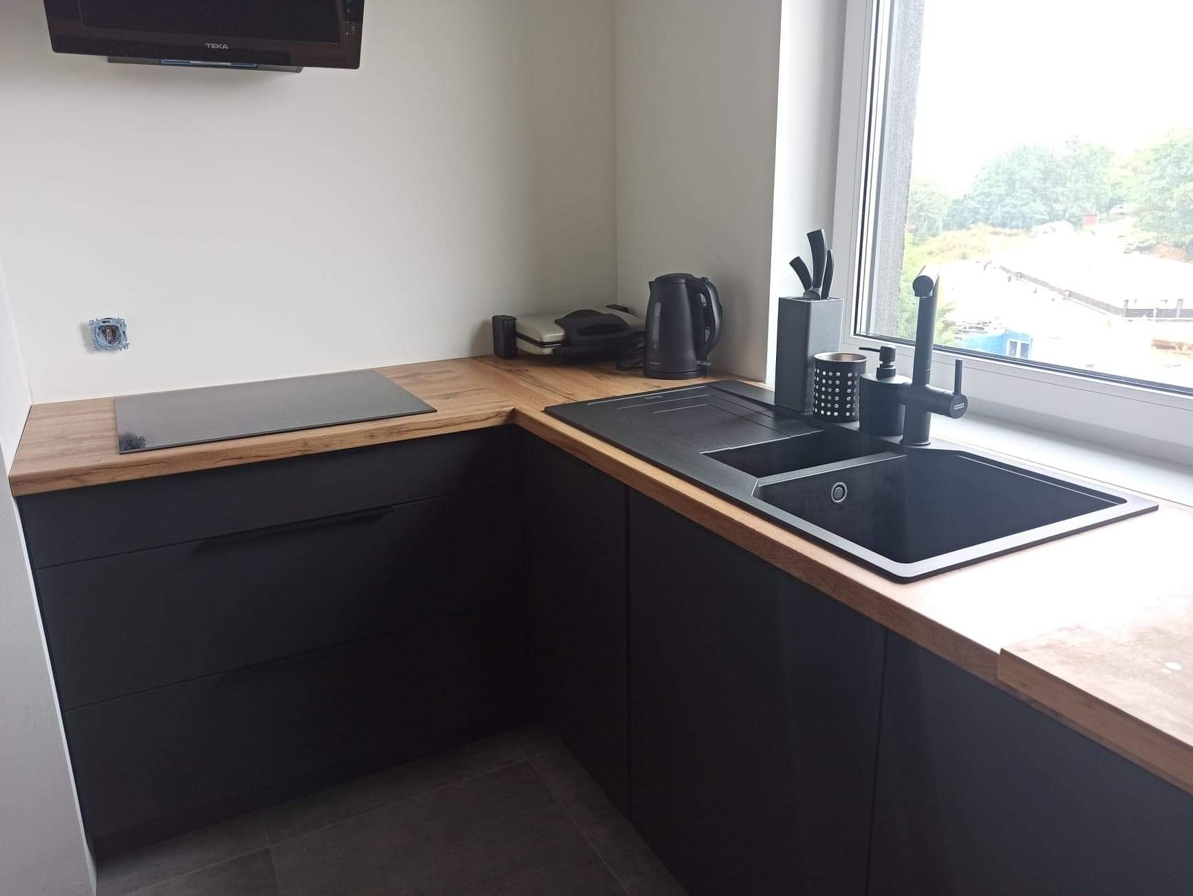 Czarna, narożna zabudowa kuchenna z drewnianym blatem, czarnym zlewem i armaturą, widok na okno i fragment płyty indukcyjnej.