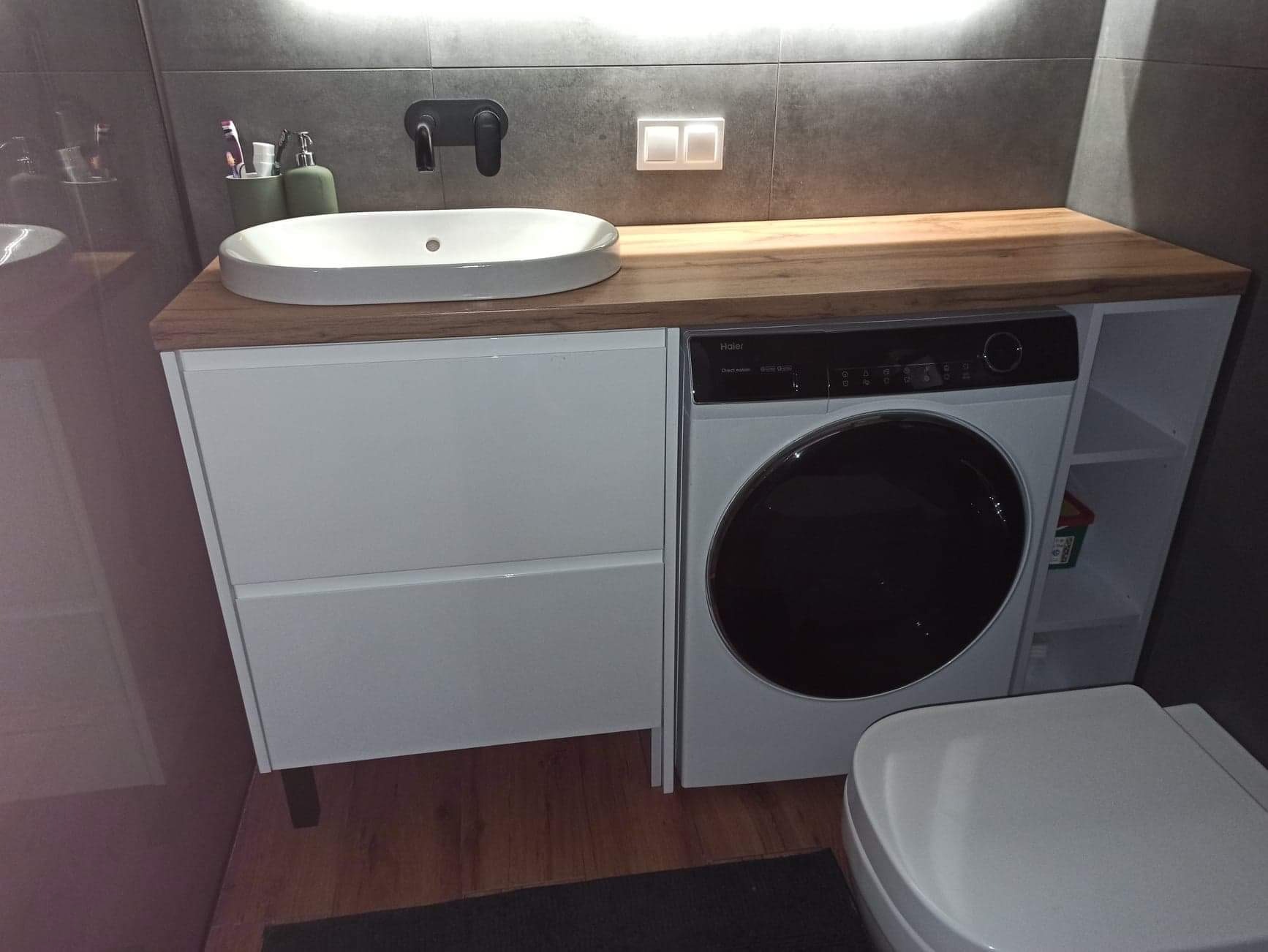 Nowoczesna łazienka z blatem drewnianym, umywalką nablatową, białą szafką z szufladami i pralką zabudowaną pod blatem, szara ściana z czarną armaturą.