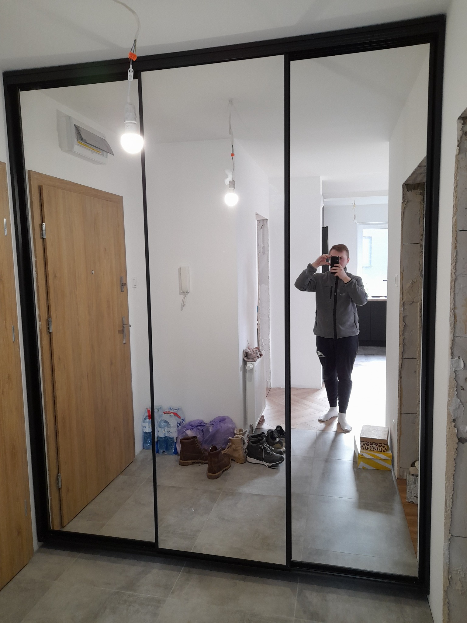 Nowoczesna szafa wnękowa z czarnymi ramami i lustrzanymi drzwiami przesuwnymi, odbijająca postać fotografa robiącego zdjęcie w korytarzu, widoczne buty i butelki z wodą przy drzwiach.