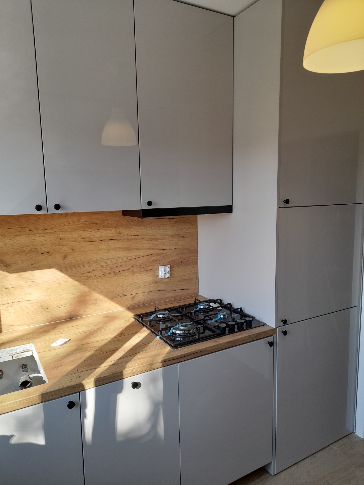 Szare, lakierowane szafki kuchenne z drewnianym blatem i płytą gazową, widoczny otwór na zlew w trakcie montażu, oświetlone popołudniowym słońcem.