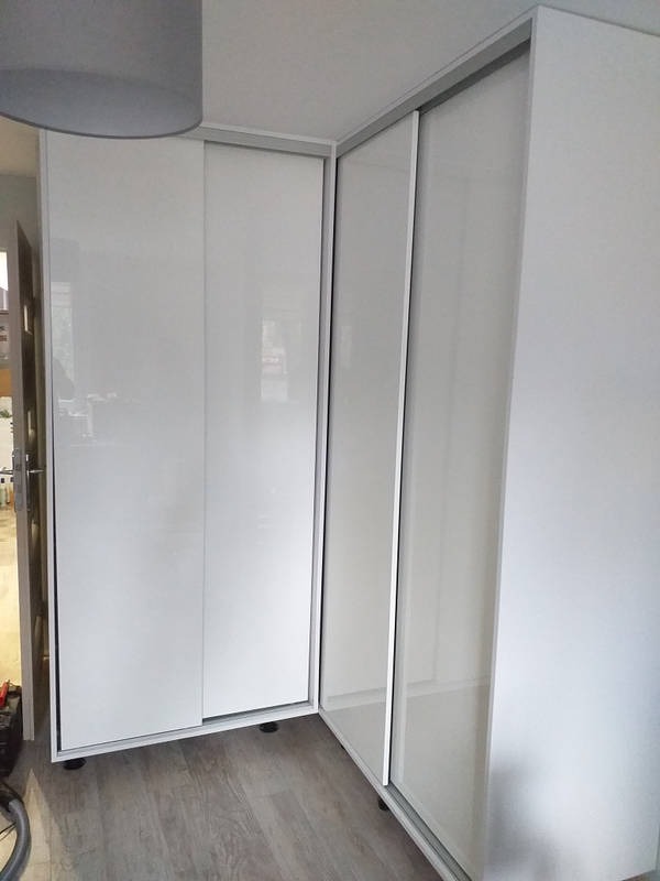 Biała szafa wnękowa z przesuwnymi, lakierowanymi drzwiami, zamontowana w rogu pomieszczenia, widoczna z boku z szarym abażurem lampy w tle.