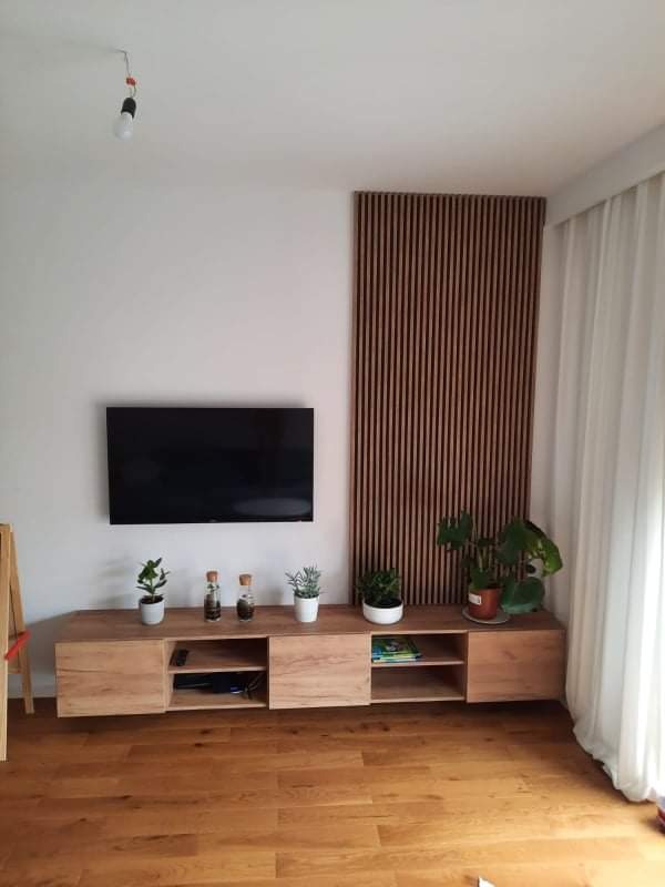 Jasna, drewniana szafka RTV z przesuwnymi drzwiczkami i otwartymi półkami, na niej rośliny doniczkowe i dekoracyjne butelki, za nią panel ścienny z drewnianych listewek, podłoga z desek w ciepłym...