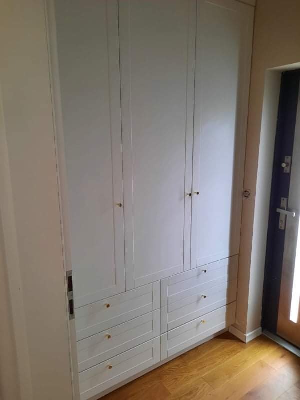 Biała szafa z czterema drzwiami i sześcioma szufladami z okrągłymi, złotymi uchwytami, wbudowana we wnękę przy drzwiach wejściowych.