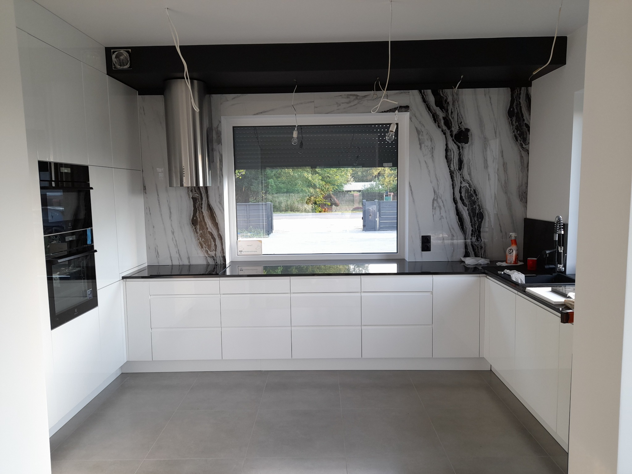 Nowoczesna, biała kuchnia z czarnym blatem i marmurowym panelem ściennym; widoczne niedokończone podłączenia elektryczne.