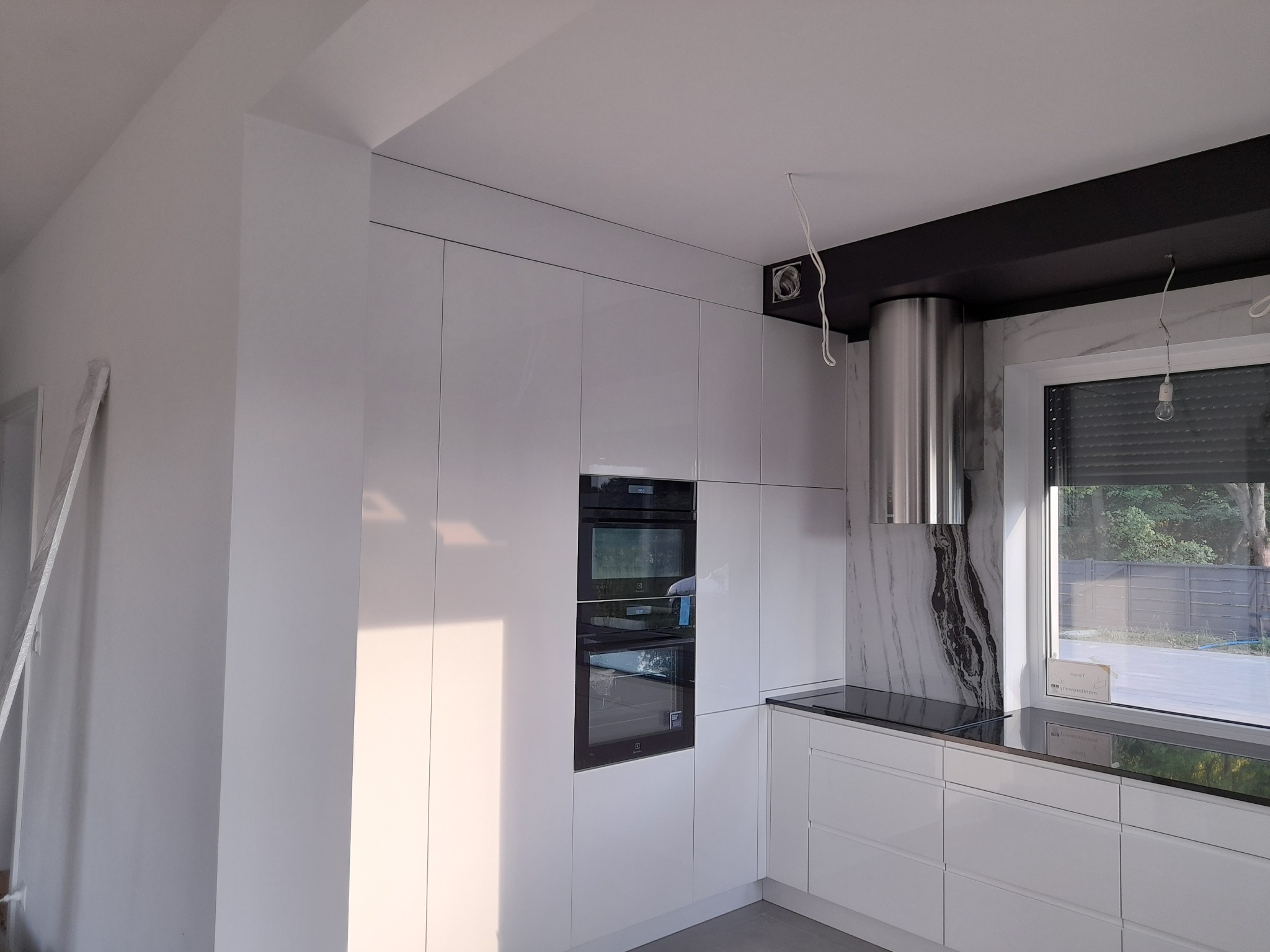 Nowoczesna kuchnia z białymi, lakierowanymi frontami, czarnym blatem i obudową okapu, w trakcie montażu widoczne kable oświetleniowe.