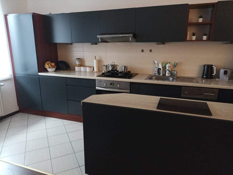 Ciemna zabudowa kuchenna z wyspą, widoczne naczynia, owoce i akcesoria kuchenne, płytki podłogowe w jasną kratę.