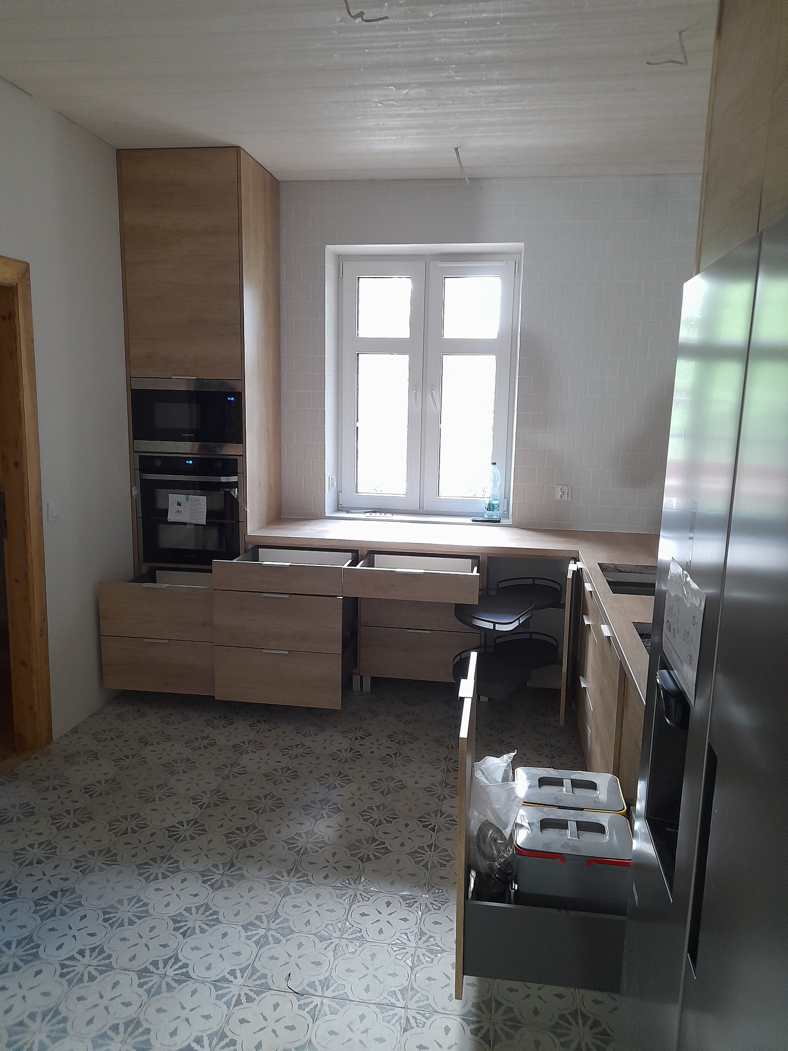 Wnętrze kuchni w trakcie montażu, z otwartymi szufladami i widocznymi elementami konstrukcyjnymi, jasne drewno frontów i wzorzysta podłoga.