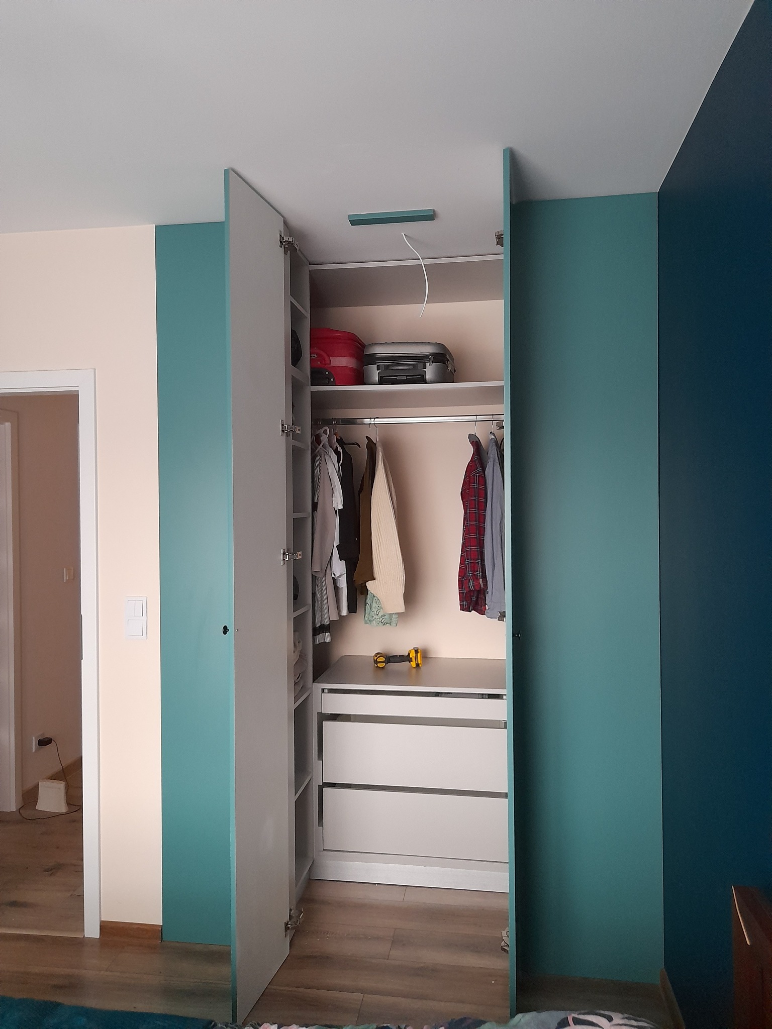 Otwarta szafa wnękowa z szarymi drzwiami i turkusowymi bokami, widoczne ubrania na wieszakach, szuflady i walizki na górnej półce, żółta miarka leży na blacie.