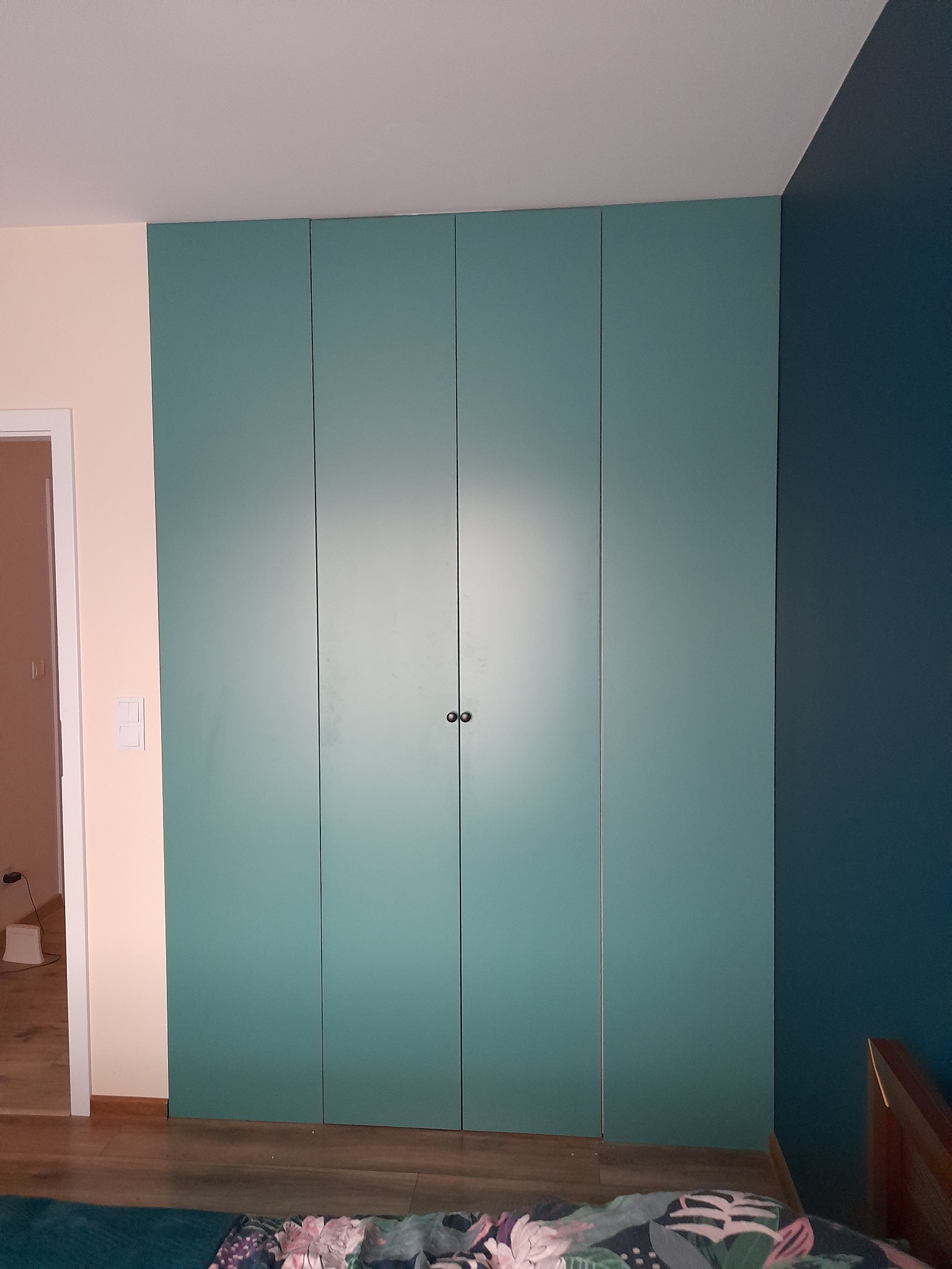 Czterodrzwiowa szafa w kolorze morskim, wkomponowana w narożnik pokoju z dwiema różnymi kolorami ścian, widoczna z perspektywy niskiego kąta.