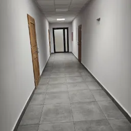 Długi korytarz z szarymi płytkami podłogowymi i białymi ścianami, z trzema drewnianymi drzwiami po lewej stronie i czarnymi drzwiami na końcu korytarza.