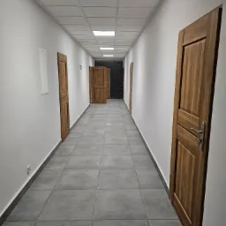 Długi korytarz biurowy z szarymi płytkami podłogowymi i białymi ścianami, z drewnianymi drzwiami po obu stronach, oświetlony panelami sufitowymi.