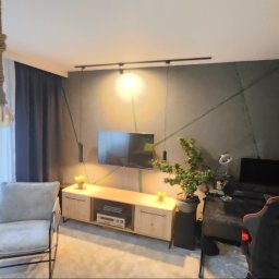 R-KWADRAT Mateusz Ryszka - Nowoczesny salon z szarą ścianą, telewizorem, drewnianą komodą i designerskim oświetleniem. Widoczny fotel i osoba w tle. Minimalistyczny wystrój wnętrza.