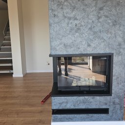 R-KWADRAT Mateusz Ryszka - Wykończony kominek z szarą, dekoracyjną strukturą na ścianie, widoczny w salonie z drewnianą podłogą. W kominku ustawione białe świece. W tle schody.