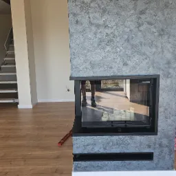 Wykończony kominek z szarą, dekoracyjną strukturą na ścianie, widoczny w salonie z drewnianą podłogą. W kominku ustawione białe świece. W tle schody.