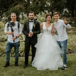 Uśmiechnięta panna młoda w białej sukni i pan młody w smokingu pozują z dwoma osobami, z których jedna trzyma kamerę na stabilizatorze, na tle zieleni.