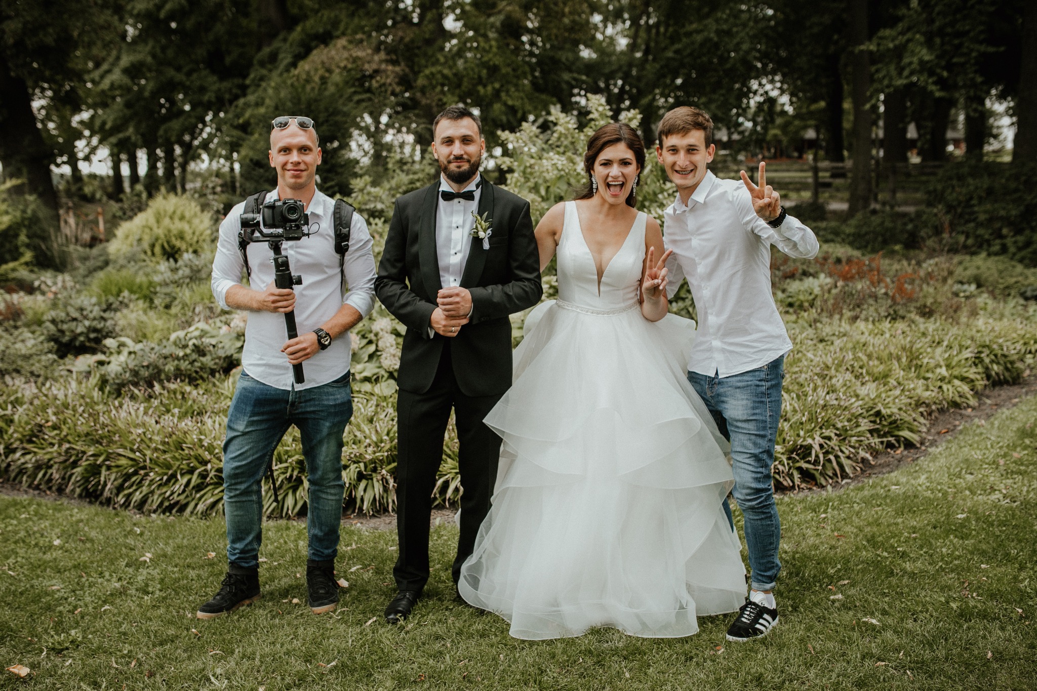 Uśmiechnięta panna młoda w białej sukni i pan młody w smokingu pozują z dwoma osobami, z których jedna trzyma kamerę na stabilizatorze, na tle zieleni.
