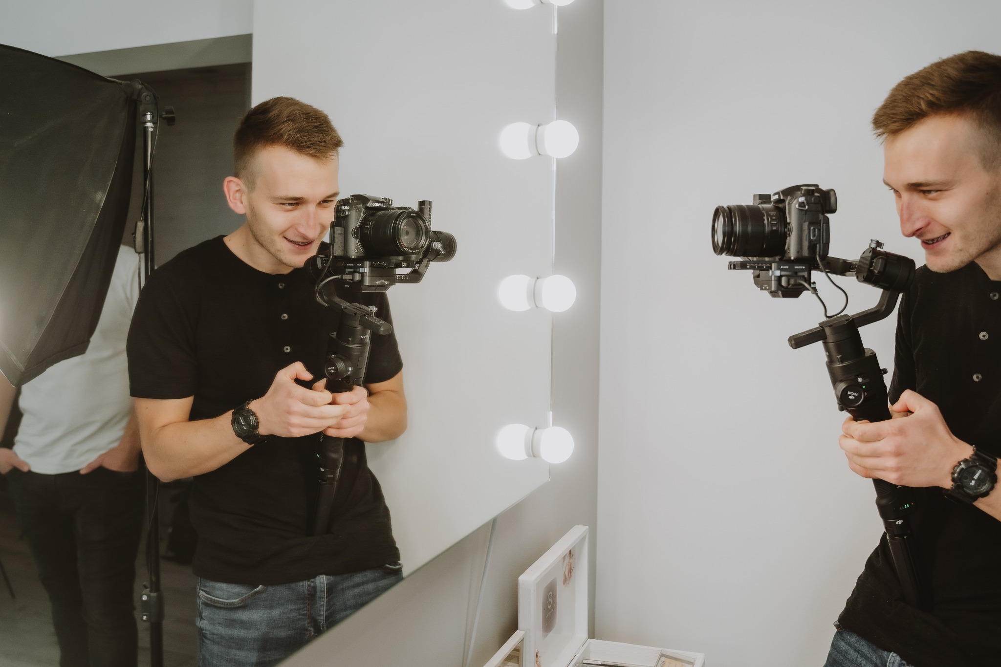 Uśmiechnięty mężczyzna z krótkimi włosami, ubrany w czarną koszulkę i jeansy, trzyma profesjonalny aparat z gimbalem przed lustrem z oświetleniem, w tle softbox.