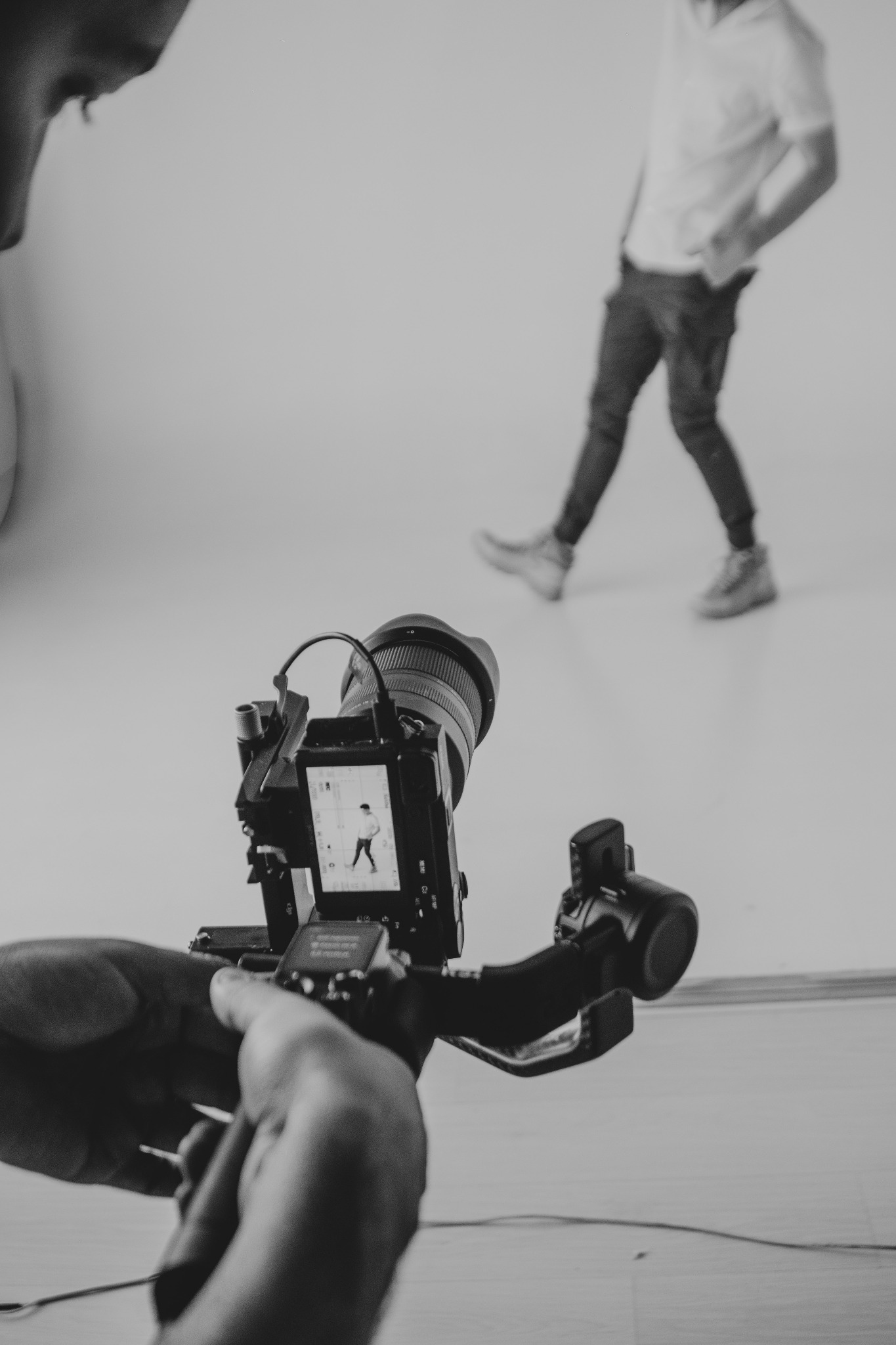 Czarno-białe ujęcie kamery na stabilizatorze, w tle rozmyta sylwetka osoby na białym tle, widoczna twarz osoby filmującej z lewej strony kadru.