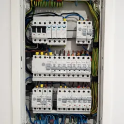 Wnętrze rozdzielnicy elektrycznej z widocznymi bezpiecznikami, przewodami i listwami zaciskowymi, zamontowanej w białej obudowie w ścianie.