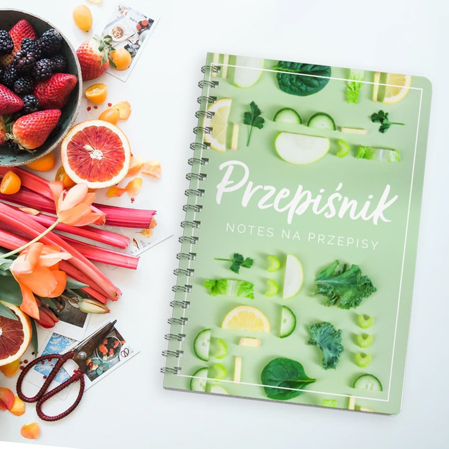Notatnik na przepisy z okładką w pastelowej zieleni, ozdobioną grafiką z warzyw i owoców, leżący obok miski z owocami leśnymi, rabarbaru i vintage nożyczek.