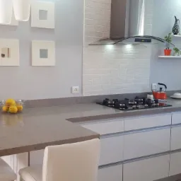 Jasna kuchnia z blatem w odcieniach szarości, minimalistyczne białe szafki, płyta indukcyjna i dekoracje na półkach.