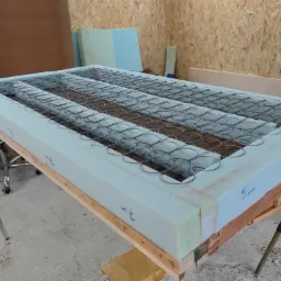 Wnętrze tapicerowanego mebla w trakcie renowacji: sprężyny bonellowe osadzone w piance, widoczne oznaczenia wymiarów na piance, drewniany szkielet na stole warsztatowym.