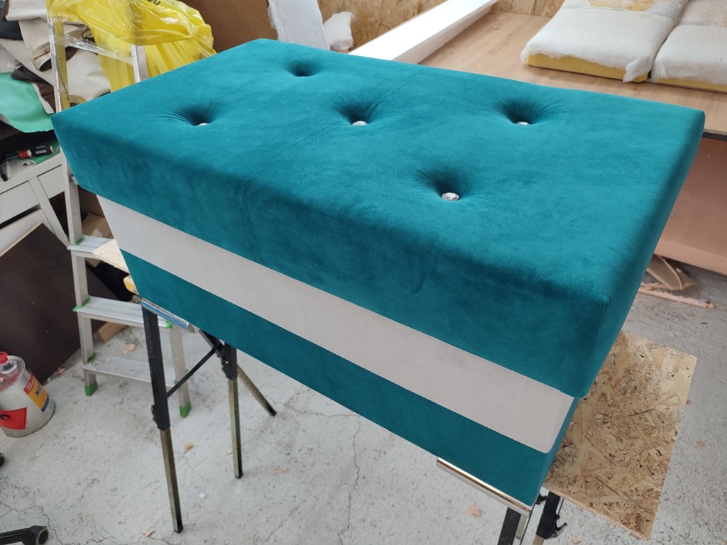 Turkusowa pufa z weluru z białym pasem, tapicerowana guzikami, na metalowych nóżkach, w warsztacie.