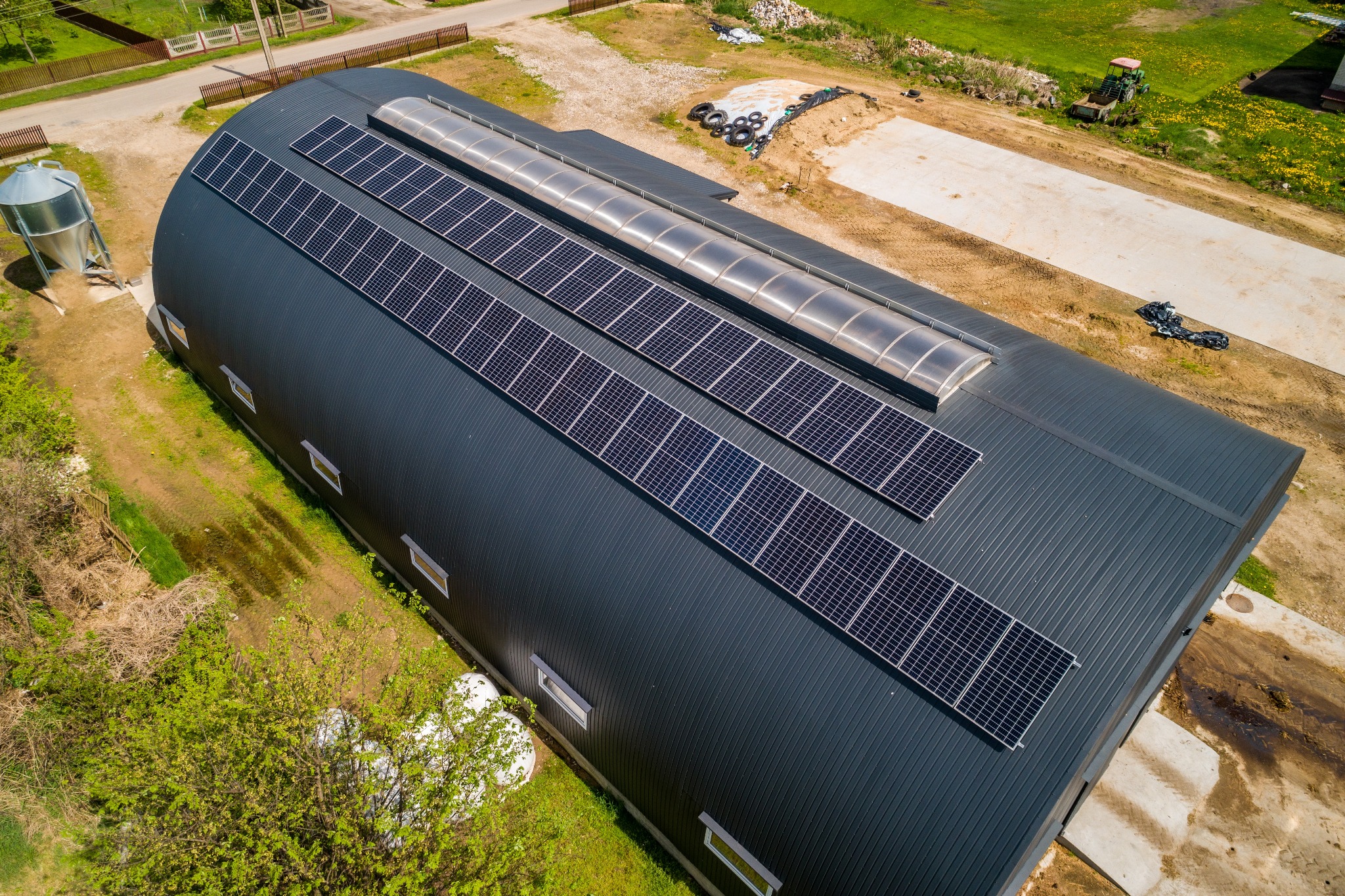 Widok z lotu ptaka na duży, łukowy budynek z ciemnoszarą blachą falistą, na dachu zamontowane panele fotowoltaiczne w trzech rzędach oraz świetliki dachowe, obok silos i teren z traktorem.