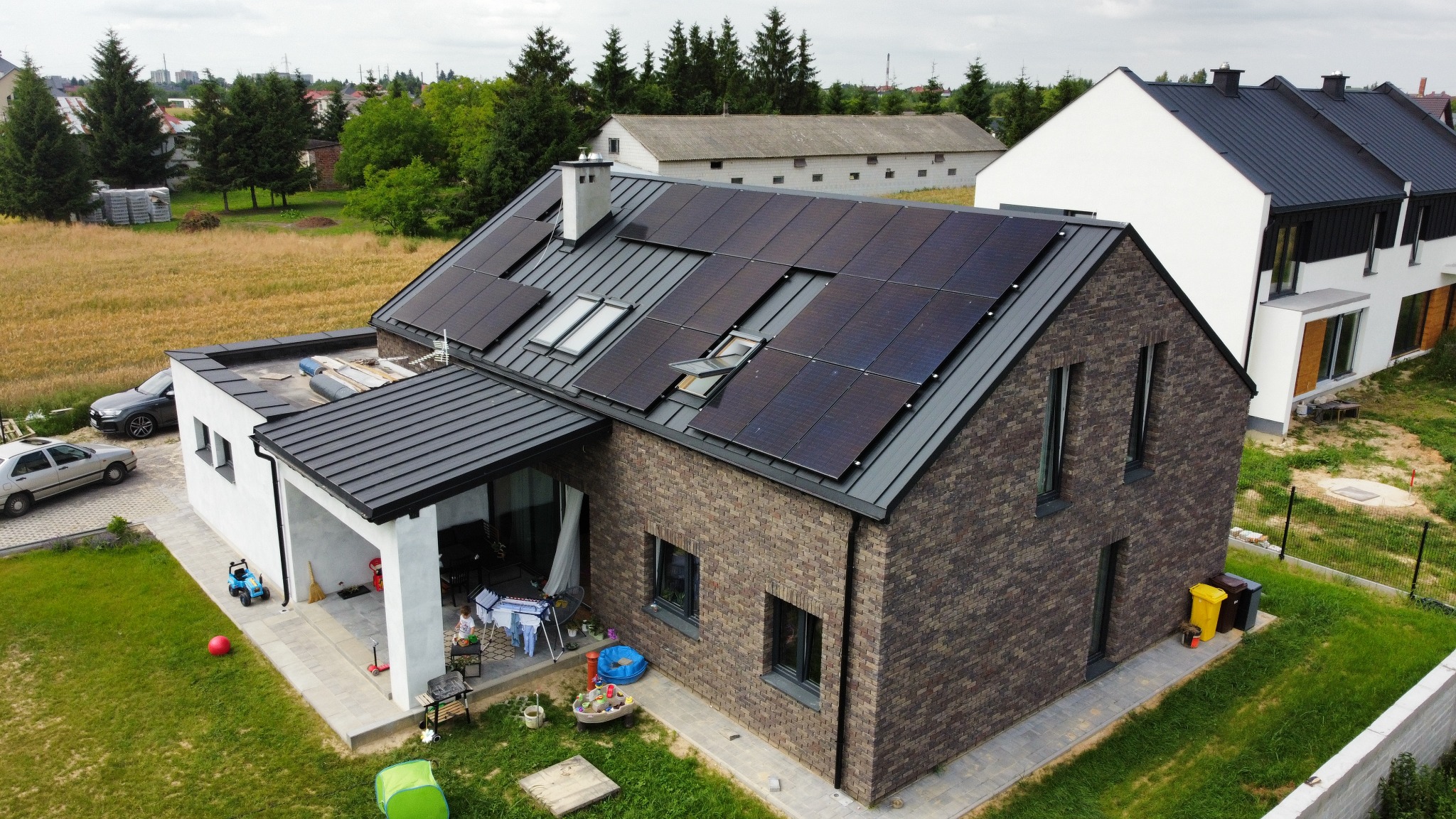 Dom jednorodzinny z brązowej cegły z panelami słonecznymi na dachu, widoczne otwarte okno dachowe, biała dobudówka z zadaszonym tarasem, na którym suszy się pranie, w tle inne budynki i pola.