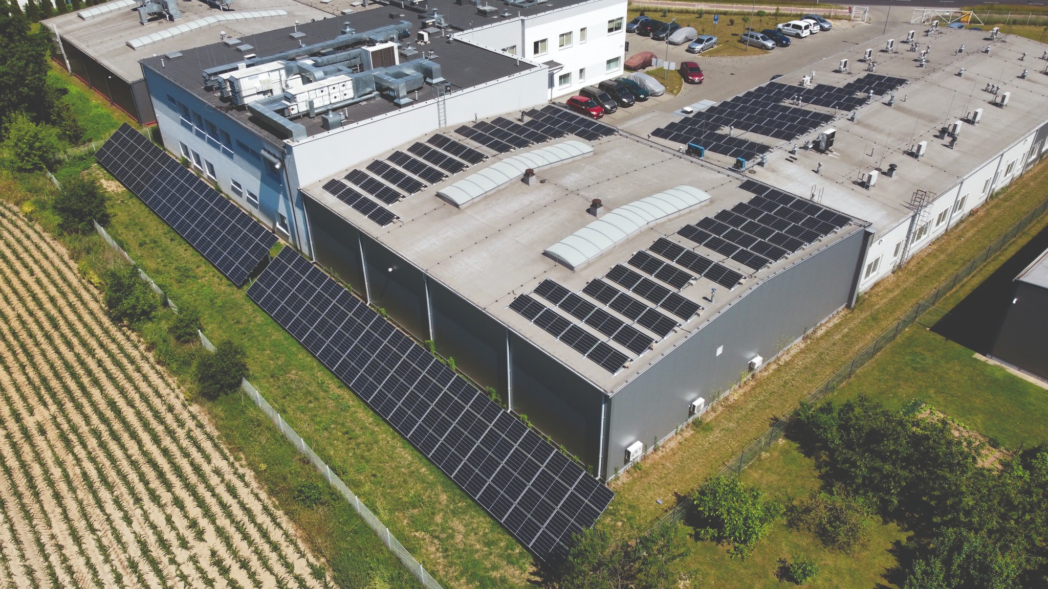 Widok z góry na budynek przemysłowy z panelami słonecznymi na dachu i gruncie, otoczony polem uprawnym i parkingiem z samochodami.