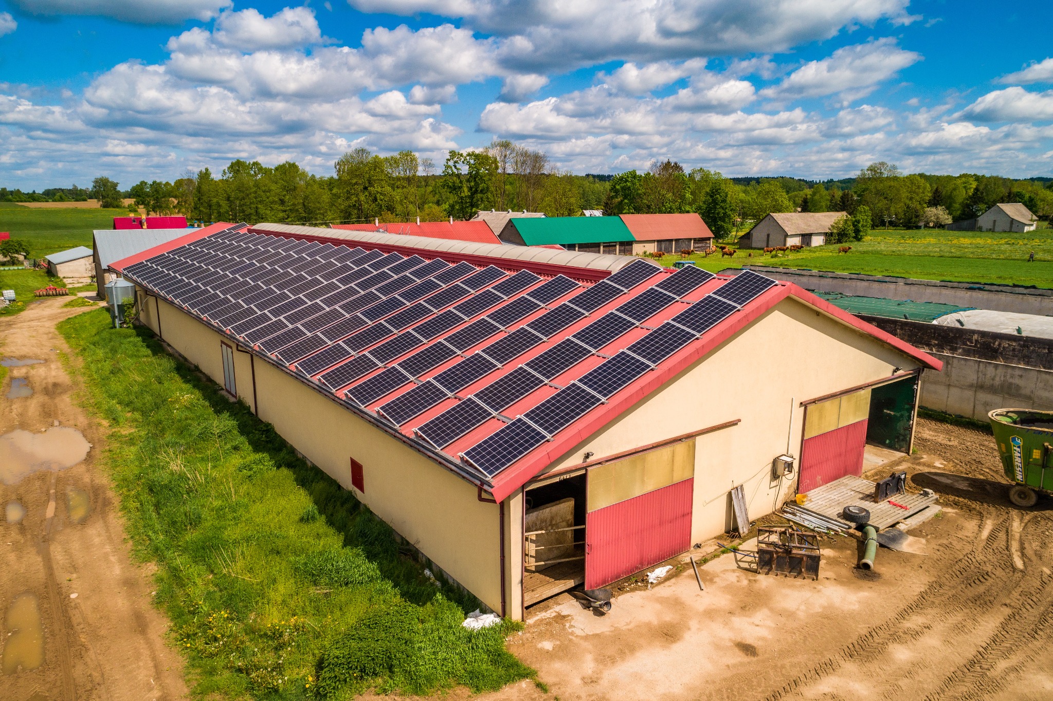 Widok z drona na duży budynek gospodarczy z panelami słonecznymi na czerwonym dachu, otoczony zielonymi polami i drzewami w słoneczny dzień.