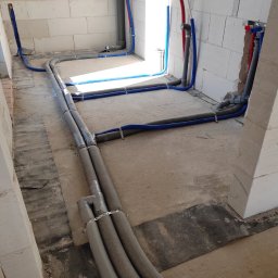 MSinstalacje Maciej Stępkowski - Instalacja wodna i kanalizacyjna w surowym stanie, rury izolowane szarą otuliną, niebieskie rury PEX mocowane do podłoża metalowymi uchwytami, widoczne przyłącza do ściany z bloczków.