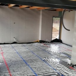 MSinstalacje Maciej Stępkowski - Rozłożona instalacja ogrzewania podłogowego z rurkami w kolorze białym, oznaczonymi liniami w kolorach czerwonym i niebieskim, na czarnej siatce w surowym wnętrzu domu w trakcie budowy.