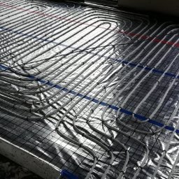 MSinstalacje Maciej Stępkowski - Rozłożone rury ogrzewania podłogowego na folii aluminiowej z siatką, widoczne niebieskie i czerwone oznaczenia, w tle okno z widokiem na plac budowy.