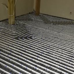 MSinstalacje Maciej Stępkowski - Białe rury ogrzewania podłogowego ułożone w pętle na czarnej siatce w surowym pomieszczeniu z drewnianą konstrukcją stropu.