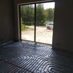 Instalacja ogrzewania podłogowego w trakcie realizacji w Łodzi, widoczne rury na folii izolacyjnej przed wylewką, z widokiem na podw&oacute;rko przez duże okno.