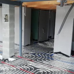 MSinstalacje Maciej Stępkowski - Białe rurki ogrzewania podłogowego ułożone na czarnej siatce, widoczne elementy konstrukcyjne budynku: drewniane belki stropowe i betonowe słupy.