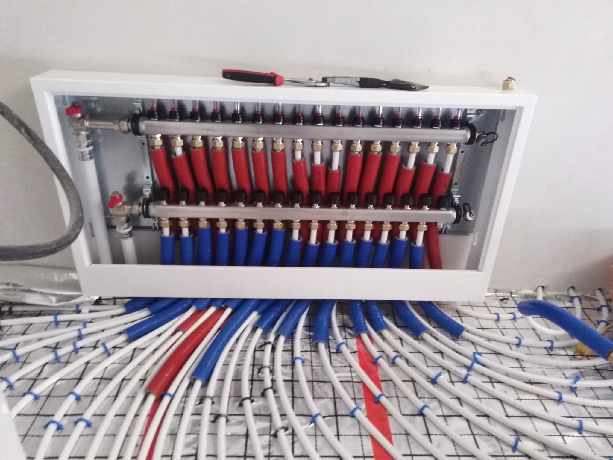 Rozdzielacz ogrzewania podłogowego z rurami w izolacji czerwonej i niebieskiej, zamocowanymi do podłoża za pomocą spinek, widoczne narzędzia na górnej półce.