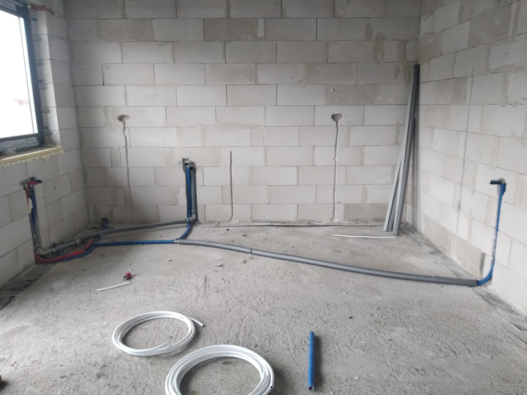 Surowy stan: instalacja wodno-kanalizacyjna w pomieszczeniu z bloczków betonowych, widoczne rury doprowadzające wodę i odpływ, oraz zwój rur do ogrzewania podłogowego leżący na posadzce.