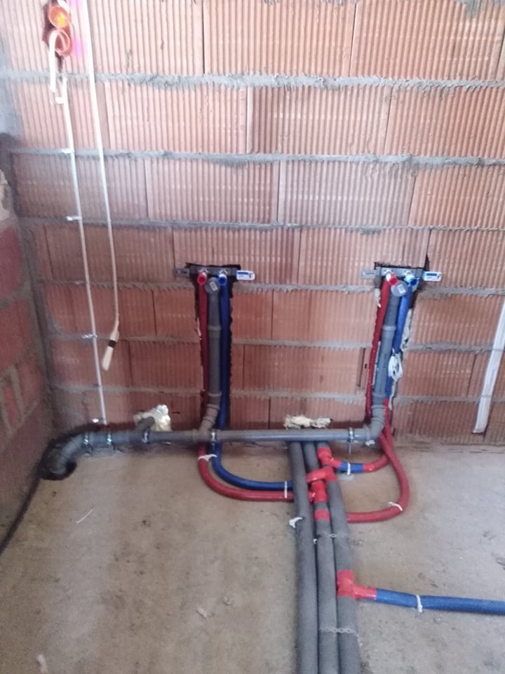 Surowe ściany z cegły dziurawki i częściowo ułożona instalacja hydrauliczna z rur szarych, niebieskich i czerwonych, prowadząca do dwóch punktów w ścianie, widoczne mocowania i izolacja rur.