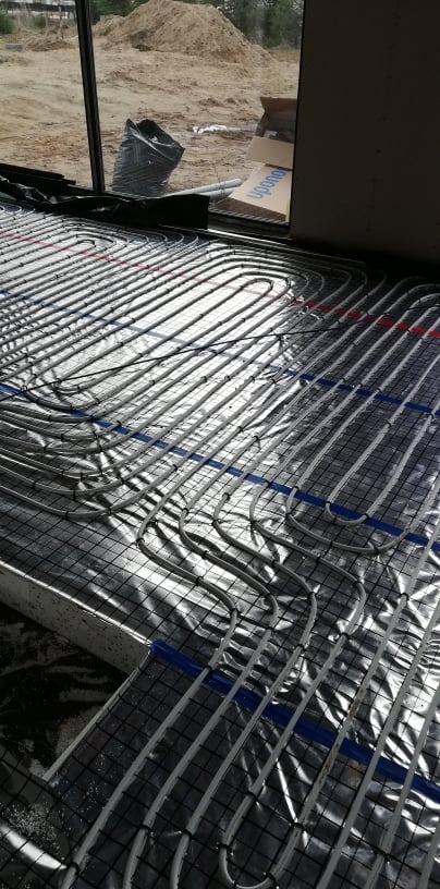 Rozłożone rury ogrzewania podłogowego na folii aluminiowej z siatką, widoczne niebieskie i czerwone oznaczenia, w tle okno z widokiem na plac budowy.