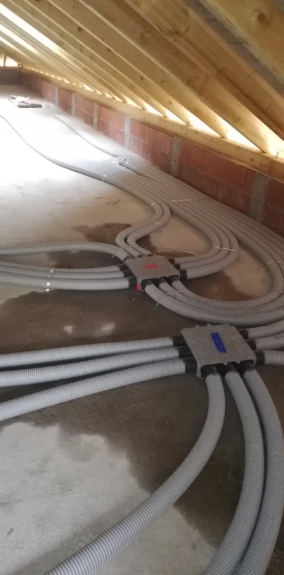 Szare, karbowane rury rekuperacji rozchodzące się z dwóch szarych skrzynek rozdzielczych na poddaszu z drewnianą więźbą dachową i fragmentem ściany z cegły.