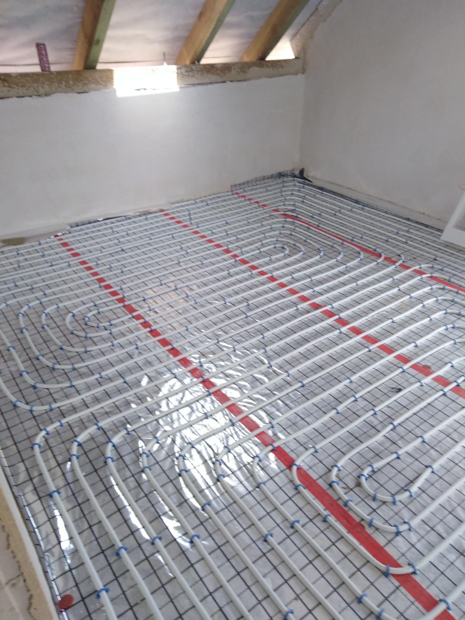 Białe rury ogrzewania podłogowego ułożone na folii z siatką w pomieszczeniu ze skośnym sufitem, widoczne drewniane elementy konstrukcyjne.